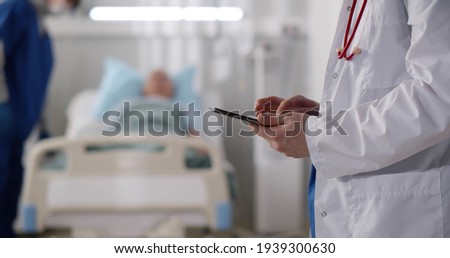Cropped shot of doctor in uniform with stethoscope using tablet pc standing in hospital ward with patient resting in bed on background. Practitioner checking patient data on digital tablet