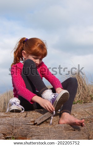little red haired girl tying up her shoelaces while sitting on a log at the beach