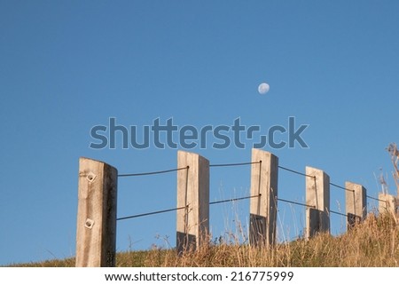 late afternoon moon rising against a blue sky with fence line