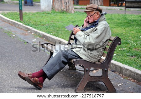 ROME, ITALY - APRIL 30, 2014: Old man sits on the bench in the park with a newspaper on his lap and speaks on the cell phone