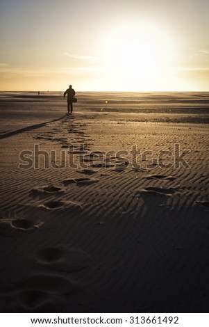 Man with suitcase walking on the beach towards the setting sun