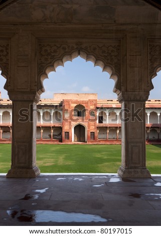 Private courtyard seen from under arches of Palace at Agra Fort in India. Green grounds, blue skies, beige arches frame red gate and white gallery.