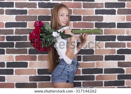 Young beautiful blond smiling girl holding roses wearing jeans shorts and white long sleeve shirt standing near the brick wall
