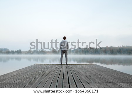 Young man standing alone on wooden footbridge and staring at lake. Thinking about life. Mist over water. Foggy air. Early chilly morning. Peaceful atmosphere in nature. Enjoying fresh air. Back view.