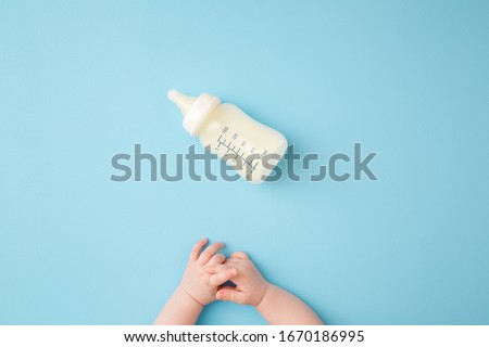 Infant hands and bottle of milk on light blue table background. Feeding time. Pastel color. Closeup. Point of view shot.
 