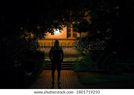 Hooded man standing on sidewalk at dark park in summer black night. Scary moment and gloomy atmosphere. Thief watching house. Back view.