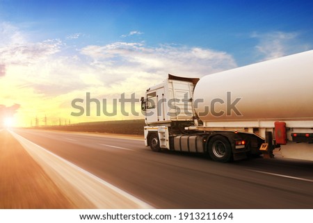 A big metal fuel tanker truck shipping fuel on the countryside road in motion against a blue sky with a sunset
