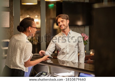 A female hotel receptionist taking the contactless payment of a customer who is checking in to the hotel, he is using his smart phone to pay.