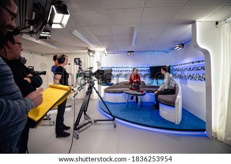 A TV show being filmed in a studio. The presenters are sitting on the studio sofa and talking to each other while the camera crew films them with film cameras.