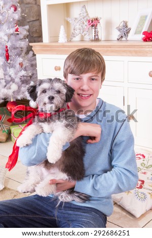 Boy holding a Puppy at Christmas Time. He is Looking at the Camera and Smiling and the Puppy has a Red Bow around its neck.