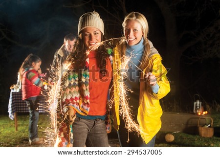 Two women are standing together smiling for the camera with lit sparklers in their hands. They are outside and there are other people in the background behind them.