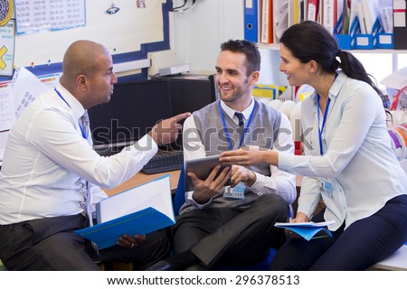 School teachers gather in a small school office for a chat. They look happy. A woman and two men group together. A man holds a digital tablet