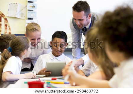 A male teacher sits supervising a group of children who are working on whiteboards and digital tablets.