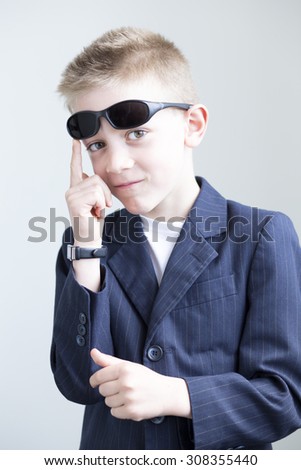 Young boy dressed up and posing like a spy. He is wearing a suit with sunglasses on his head and a formal watch. He is miming a gun with his hands.