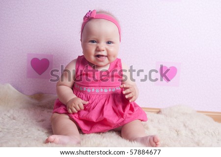 Smiling little baby girl sitting on the carpet at the pink wall with hearts in pink headband and dress