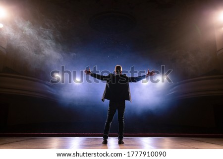 Showman. Middle Age Male entertainer, presenter or actor on stage. Arms to sides, smoke on background of spotlight. Rear view portrait of a male public speaker speaking at the microphone, pointing