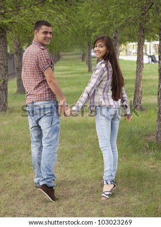 Girl with a guy in a checkered shirt and blue jeans walking in the park among the rows of trees, holding hands and looking at each other