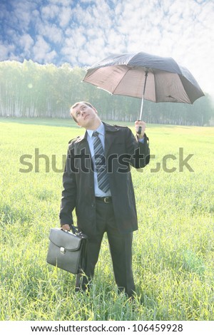 A unique view of a businessman, standing on meadow , holding an umbrella on a bright sunny day, reaching out with his hand as it to check to see if it is raining.