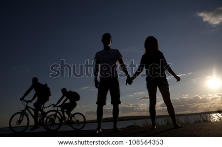 silhouettes of four young people - the first pair is on the road and holding hands - second ride bicycles