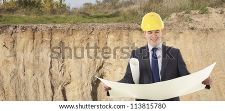 engineer in a helmet with paper plans of buildings in the trench at the site of the future foundation