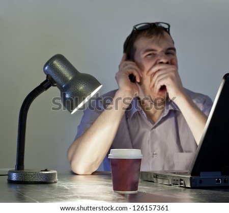 man in evening light, table lamp yawns at his desk in front of a black laptop