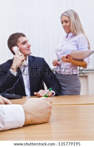 Happy businesswoman on phone call, smiling, gesturing with pen handheld, busy office in background.  writing hand with pen in focus in the foreground