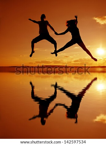 Silhouettes of couple jumping on yellow sunset background with reflection on water