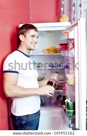 Portrait of young caucasian man looking at someone against different healthy food inside fridge and holding in hands glasses bouttle with alcohol