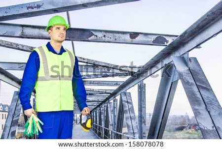 portrait of serios young builder in green and blue uniform , on old rusty metal industrial  bridge background