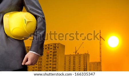 adult engineer or inspector hand holding yellow plastic helmet for workers security over highrise buildings construction cranes on background evening sunset sky Crane lifts load background No face