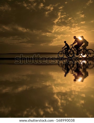 Image of sporty company three friends on bicycles outdoors against sunset. Silhouette A lot phases of motion of a single cyclist along the shoreline coast Reflection on water Space for inscription