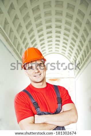 Portrait of young adult worker in blue and red uniform and security orange helmet standing above green arch building in perspective background