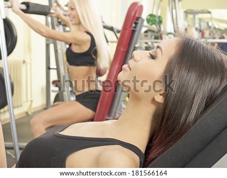 Portrait of two young adult Girls do exercise for legs and hands. in fitness gym on mirror with reflection and window background 2 woman with long blond and brunette hair sitting smiling relaxed face
