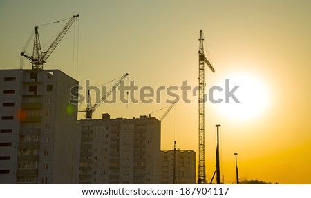 Background of Construction site with home silhouette manual worker in helmet disengages from the cargo hook on the construction crane slings on yellow sun set cloudy sky with rays light backdrop