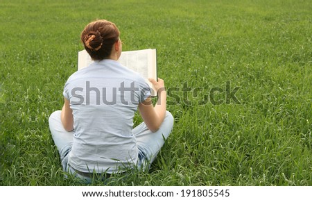 Back view young adult Girl sitting on fresh green grass background with big book Woman wearing casual blue dress Copy space for inscription