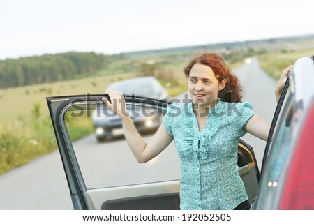 Portrait of Pretty girl is standing near the modern car against driving auto. Focus point on face Young adult traveling redhead woman based on door automobile on green grass field, forest background