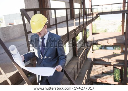 Portrait of young adult caucasian male architect in yellow hardhat looking paper plan blueprint and writing with pen documents. Horizontal shape Well dressed engineer sitting on metal rusty ladder
