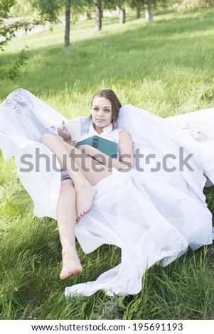 beautiful young adult caucasian girl laying with green book, has a rest in park cafe with cup caffee on fresh green grass and blooming tree background Slim female sit on white armchair and dreaming