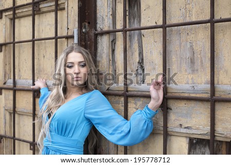 Portrait of cute Young adult woman holding on rusty metal lattice near old old retro yellow wooden door background  Sad caucasian girl wearing blue dress and looking down