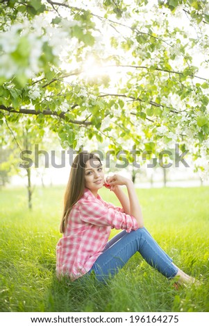 Portrait of nice dreamy girl sitting down under blooming flowers on apple tree on fresh green grass in spring garden background , sun shine in blossom, beauty of nature and woman concept