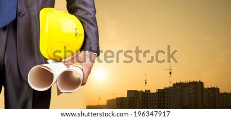 engineer hand holding yellow helmet and blueprint on background of new highrise apartment buildings and construction cranes on background of evening sunset cloudy sky Silhouette Crane lifts load