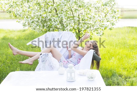 beautiful young girl sitting with cup of coffee and white sweat in park cafe  near decor table on green grass and blooming tree background female sit on white armchair Copy space for inscription