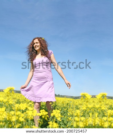 Beautiful caucasian young adult redhead curly woman in short purple dress standing on summer yellow green flower rape field outdoors at sunny day Full length Copy space for inscription