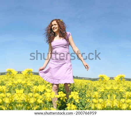 Beautiful caucasian young adult redhead curly woman in short purple dress standing on summer yellow green flower rape field outdoors at sunny day Full length  Copy space for inscription