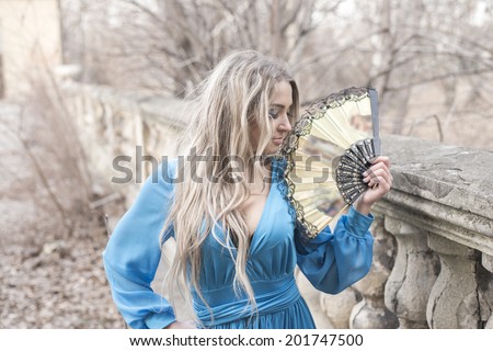 Portrait of young adult woman holding in hands old retro vintage fan Girl face with  smile on her face Cute beautiful blond female with long hair looking up and wearing stylish sexy blue dress.