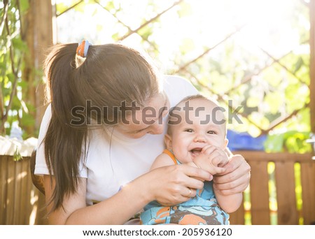 Portrait of happy little asian mom kissing son outdoors on green summer garden with fresh grass background Cute latin hispanic mother embracing baby boy looking at camera