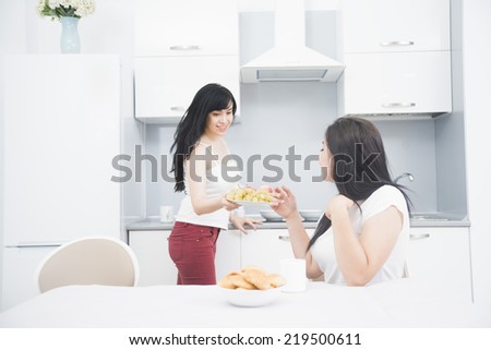 Two  brunette girls friend drinking coffee or green tea talking and looking at camera Couple of woman eating fruit - apple on plate and cookie holding in hand on kitchen interior background