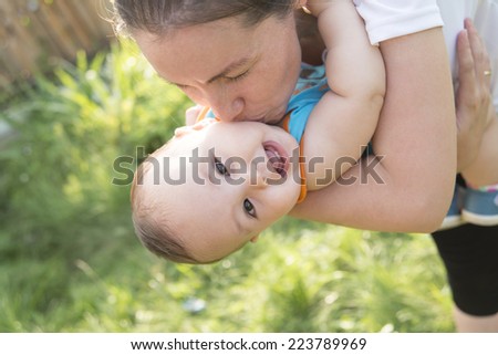 Portrait of happy little asian mom kissing and shake son outdoors on green summer garden with fresh green grass background Cute latin hispanic mother embracing baby boy looking at camera