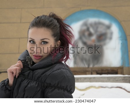 Outdoor portrait of beautiful cute asian girl  looking at camera against yellow wall with painting owl Brunette woman wearing black down jacket with big art metal earrings