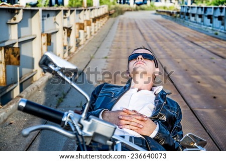 Romantic portrait resting relax handsome biker man in sunglasses sits on a bike on a sunset on old rusty metal bridge in city against house and sunny sky background Empty copy space for inscription